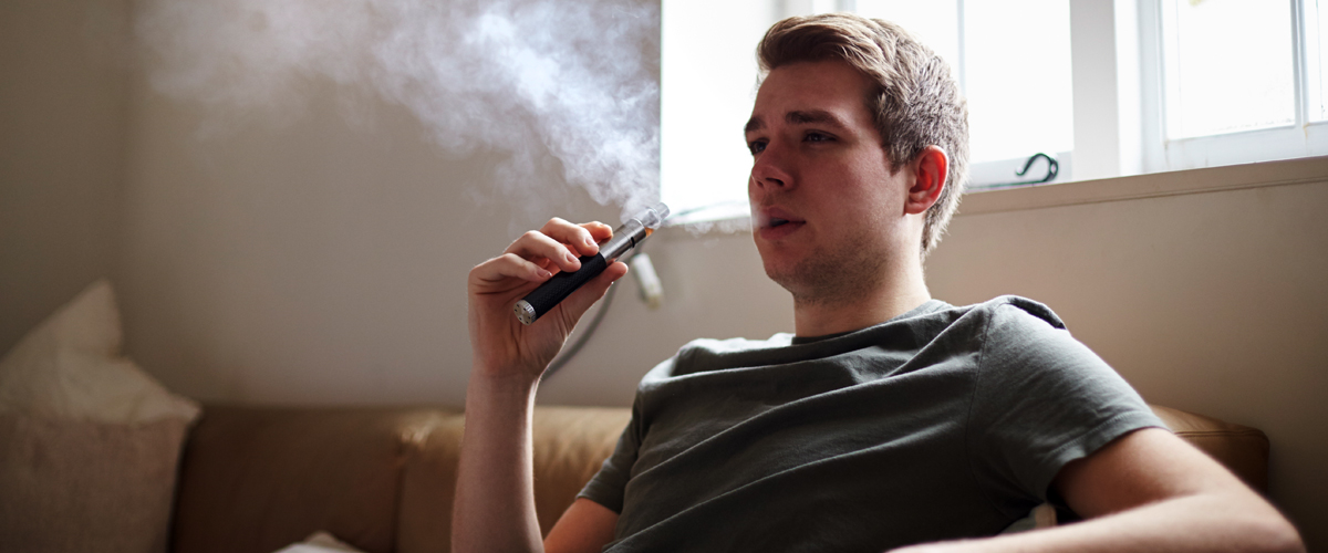 Young adult sitting on a couch, holding an e-cigarette and exhaling smoke