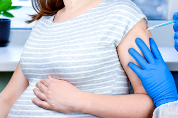 Blue gloved hands give a covid vaccine during pregnancy to a pregnant person in a striped shirt
