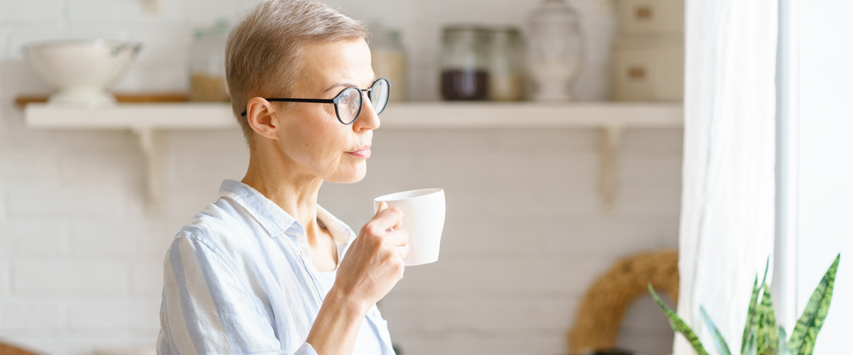 Menopause and brain: Woman holding coffee cup and looking out window with serious expression.