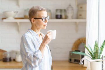 Menopause and brain health: Woman holding coffee cup and looing out window with serious expression.