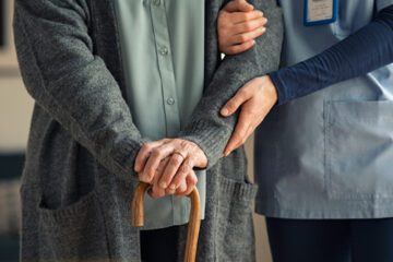 A caregiver holding onto a person with a cane.