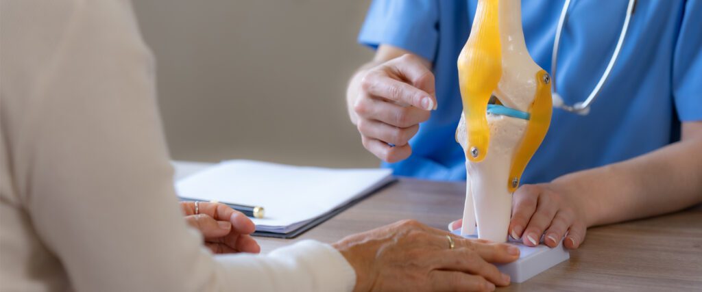A doctor points to a model of a knee joint, discussing joint replacement surgery.