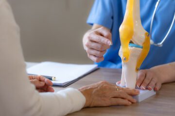 A doctor points to a model of a knee joint, discussing joint replacement surgery.