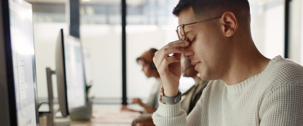Man sitting at desk with eyeglasses, holding hand to temple