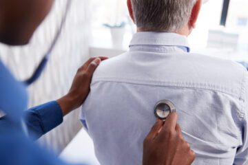 A medical professional holds a stethoscope up to a person's back to listen to their lungs.